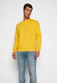 Gele sweatshirt met een ronde hals, verlaagde schouders en geribde manchetten, gecombineerd met lichtblauwe jeans met een getailleerd ontwerp.