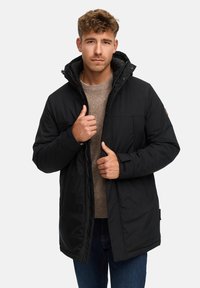 Giubbotto imbottito nero con cappuccio, tasche frontali e chiusura con zip. Il modello indossa un maglione color cammello sottostante con una finitura testurizzata.