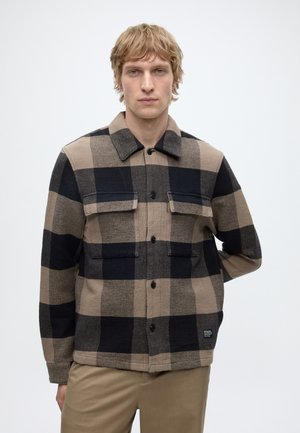 Young man with blonde hair wearing a beige and black checkered button-up jacket and beige pants against a plain light background.