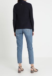 Frau trägt einen schwarzen ribbeligen Pullover, hellblaue hochgeschnittene Jeans und graue Blockabsatzsandalen mit Knöchelriemen, steht mit dem Rücken zur Kamera.