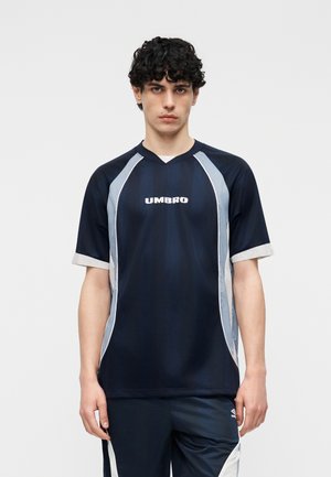 Νεαρός άνδρας με σγουρά μαλλιά, φορώντας αθλητική φανέλα Umbro σε μπλε ναυτικό και λευκό και αντίστοιχο σορτς, στέκεται με το σώμα του προς τα εμπρός μπροστά σε απλό φόντο.