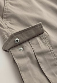 Beżowy materiałowy rękaw z klapą zabezpieczoną dwoma metalowymi zatrzaskami, pokazujący szczegółowe przeszycia i złożony mankiet.