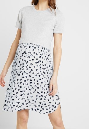 Kobieta w jasnoszarej bluzce z krótkim rękawem i przymocowaną białą spódnicą z czarnym kwiatowym wzorem, stojąca na jednolitym tle.