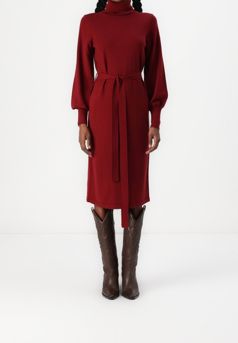 Femme portant une robe midi rouge à manches longues ceinturée, associée à des bottes en cuir marron montant aux genoux, debout devant un fond blanc uni.