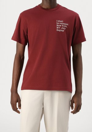Mężczyzna ubrany w bordowy t-shirt z krótkim rękawem z białym napisem na piersi: „I shall be released New York, NY - July Replay”