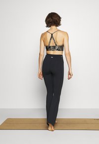 Kvinna står barfota på en yogamatta, iförd svarta tights och en mönstrad sport-bh, vänd bort mot en helt vit bakgrund.