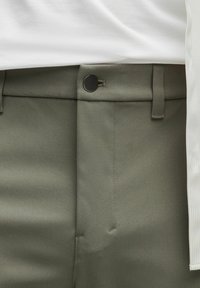 Oliwkowe spodnie z gładką fakturą, z czarnym guzikiem i paskiem, w połączeniu z białą koszulą, częściowo widoczną.