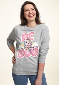 Sudadera gris con un gráfico colorido de "SE TÚ MISMO", que presenta un oso de dibujos animados y un arcoíris. Tejido suave, cuello redondo y puños de canalé.