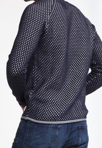 Pull bleu marine à motifs avec un design en maille triangulaire et des poignets côtelés. Comprend une bande blanche à l'ourlet et un tissu doux et texturé.