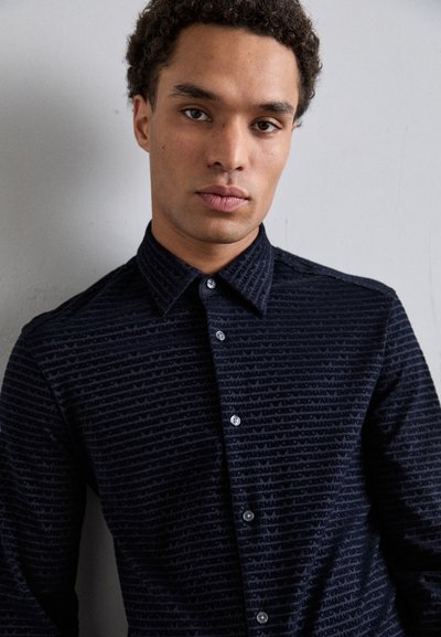 Camicia button-up blu scuro con un motivo testurizzato che presenta loghi ripetuti, colletto aperto e maniche lunghe. Chiusura standard con bottoni.