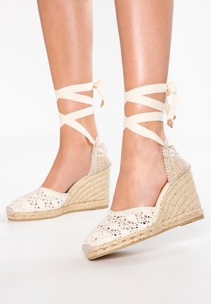 Białe koronkowe espadryle na koturnie z plecionym jutowym obcasem i beżowymi sznurówkami na kostkach. Charakteryzują się otwartym noskiem i teksturowanym tyłem dla lepszego wsparcia.