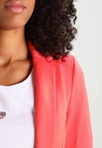 Vrouw met krullend donker haar, gekleed in een wit shirt en een koraalroze blazer, met zichtbare lippen, nek en schouder tegen een neutrale achtergrond.