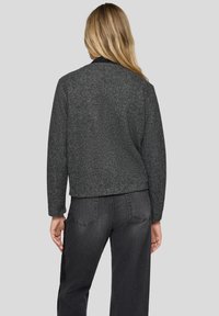 Graue, strukturierte Jacke mit abgerundetem Saum und schwarzem Kragen, kombiniert mit dunklen Jeans. Die Rückansicht zeigt eine lockere Passform und Ärmellänge.