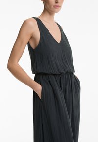 Črna enodelna obleka brez rokavov z V-izrezom, teksturirana tkanina, elastičen pas in stranske žepke, tekoč dizajn s mehkimi gubami.