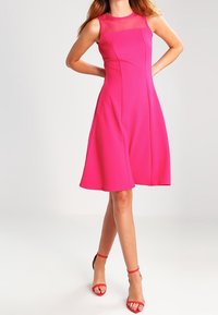 Kvinna klädd i en ärmlös ljusrosa A-linjeformad klänning, matchad med röda högklackade sandaler med ankelsnörning, står med händerna bakom ryggen.