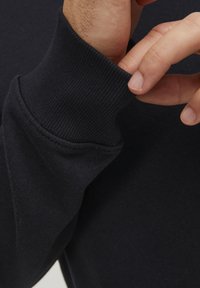 Sudadera de algodón negra con puños acanalados. Primer plano de una mano ajustando el puño, destacando la textura y el detalle de la costura.