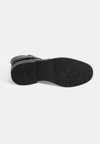 Bottine en cuir noir avec une semelle en caoutchouc texturé. Dotée d'un talon plat, d'un bout arrondi et d'un simple accent de sangle. La semelle a une adhérence à motif.