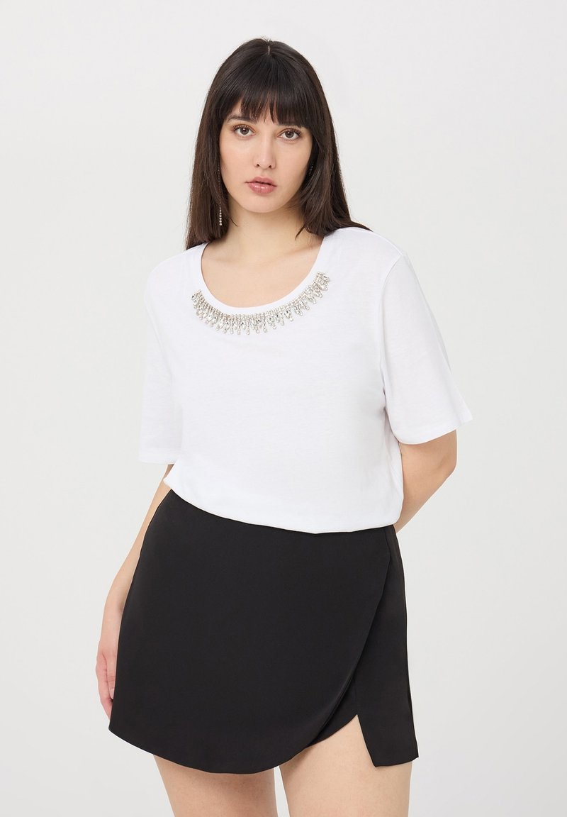 Biała koszulka z krótkim rękawem, o okrągłym dekolcie, z ozdobnym srebrnym koralikowym detalem przy kołnierzu, zestawiona z czarną spódnicą w fasonie kopertowym.