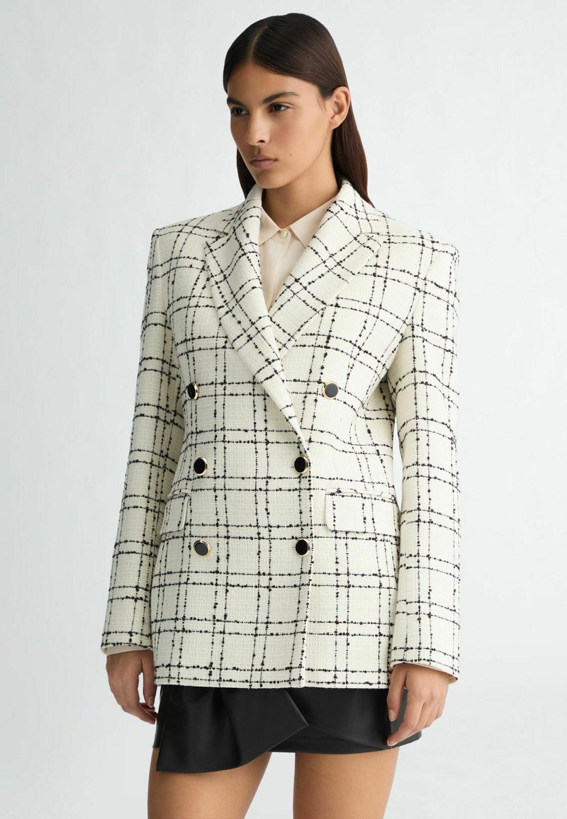 Blazer croisé en tweed crème avec un motif grille noir. Doté de boutons dorés et d'une coupe structurée, associé à une jupe noire.