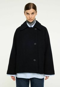 Svart oversized ullkappa med bred krage, knappstängningar och långa ärmar, lager på en blå skjorta. Texturen verkar mjuk.