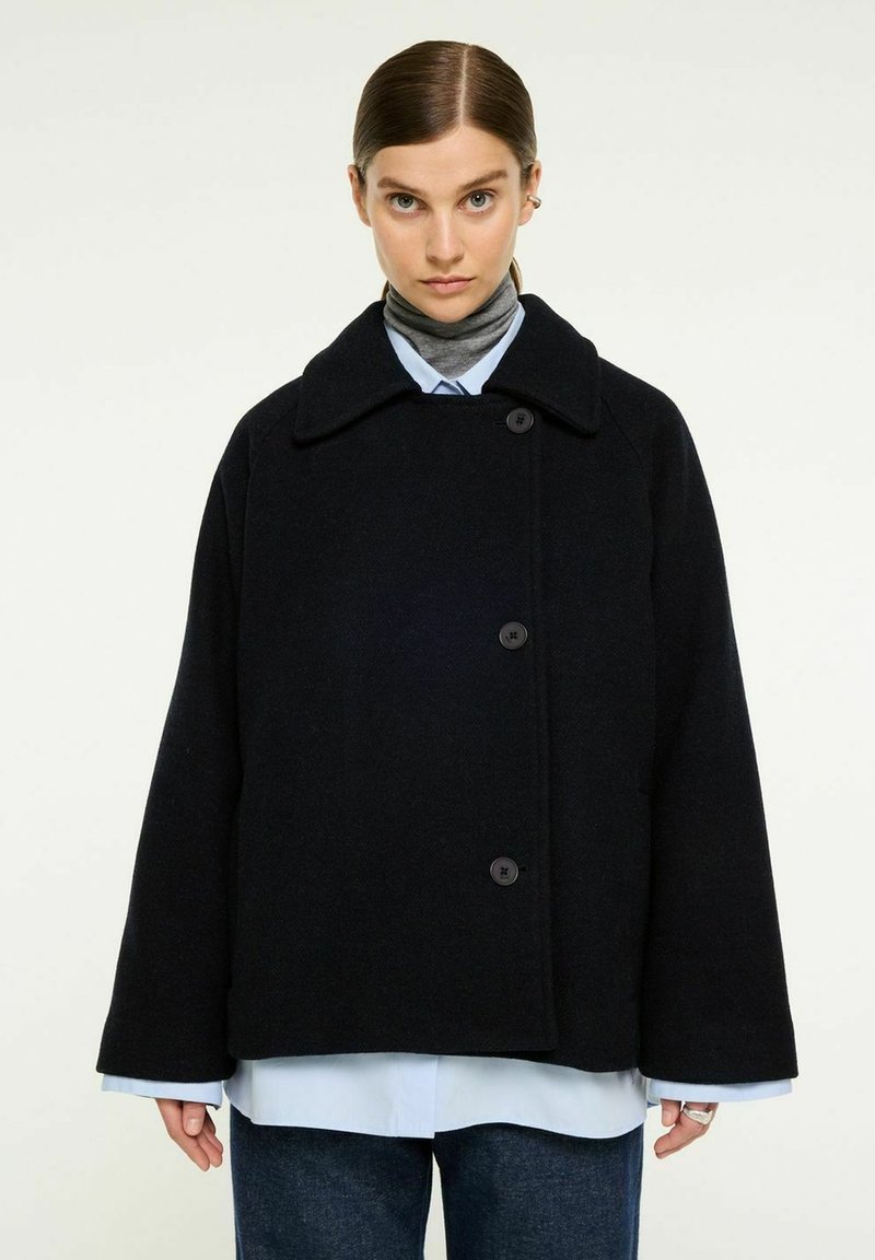 Svart oversized ullkappa med bred krage, knappstängningar och långa ärmar, lager på en blå skjorta. Texturen verkar mjuk.