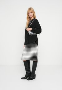Chemise noire à manches longues, robe midi rayée noir et blanc, bottes à talons hautes jusqu'au genou noires, et sac argenté au design simple.
