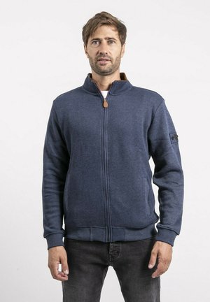 Homme aux cheveux bruns courts et à la barbe, portant un pull zippé bleu marine et un jean foncé, debout devant un fond blanc uni.