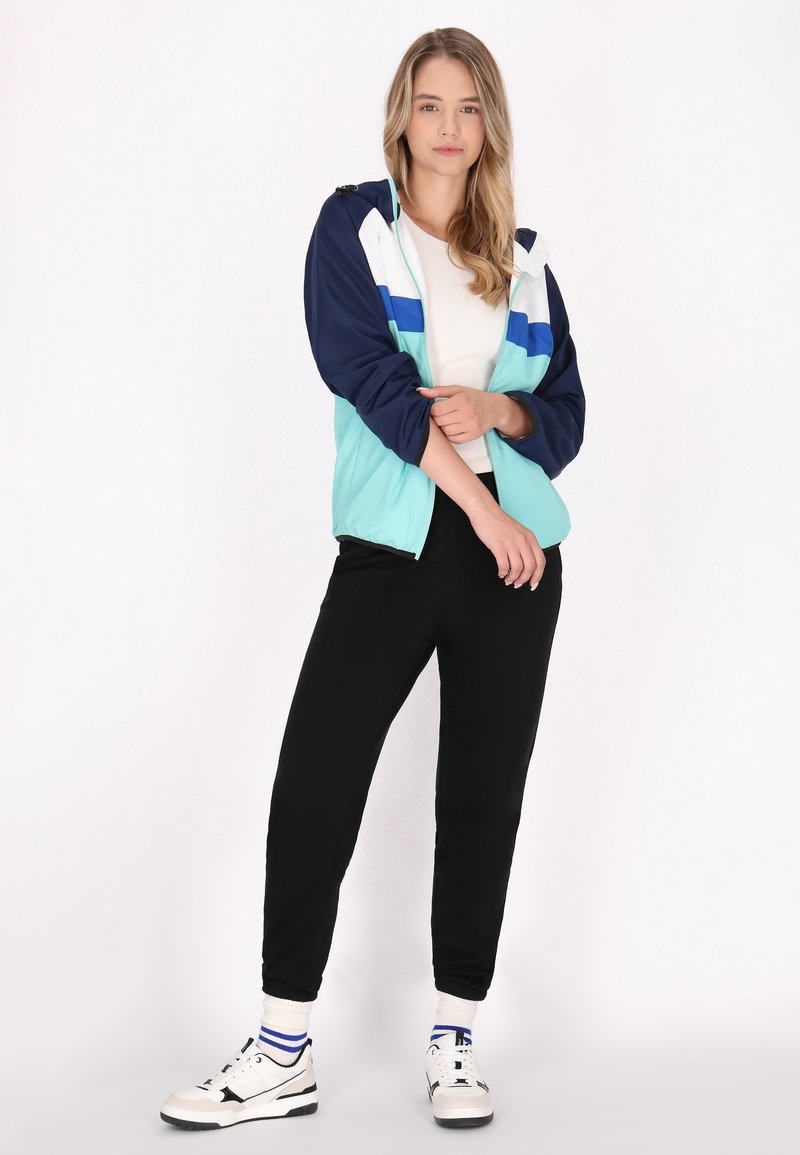 Farverig zip-up vindjakke i teal, navy og hvid med et stribet design. Sammen med sorte joggingbukser og hvide sneakers med blå accenter.