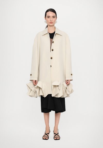 Beige coat with a collared neckline, front button closure, and ruffled hem. Worn over a black layered skirt and paired with strappy sandals.