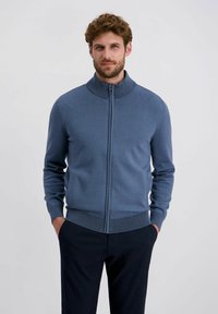 Dunkelblauer, Reißverschluss-Pullover mit hohem Kragen sowie gerippten Bündchen und Saum. Stricktextur, figurbetonter Schnitt; über dunklen Hosen getragen.