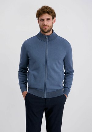 Maglione zip-up blu scuro con colletto alto e polsini e orlo a costine. Texture lavorata a maglia, design aderente; indossato sopra pantaloni scuri.