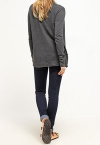 Ciemnoszary top z długim rękawem i okrągłym dekoltem, luźny krój, boczne kieszenie, zestawiony z ciemnymi dżinsami i beżowymi sandałami, widok od tyłu.