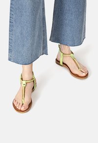 Green leather thong sandals feature braided detailing and a flat brown sole. The model wears wide blue denim pants.