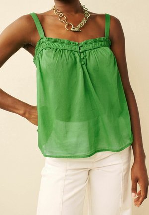 Person wearing a sleeveless green blouse with buttons and a chunky gold chain necklace, paired with off-white pants.