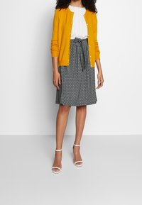 Gul knappad cardigan över en vit blus, kombinerad med en mönstrad svart kjol och vita klacksandaler.