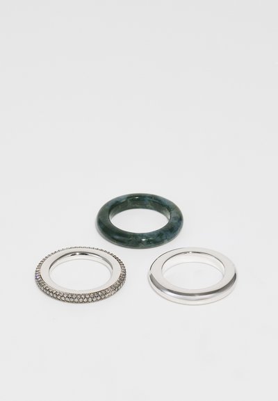 Tres anillos expuestos: un anillo de plata con piedras transparentes, una banda de jade verde, y una banda de plata lisa. Diferentes formas y texturas.