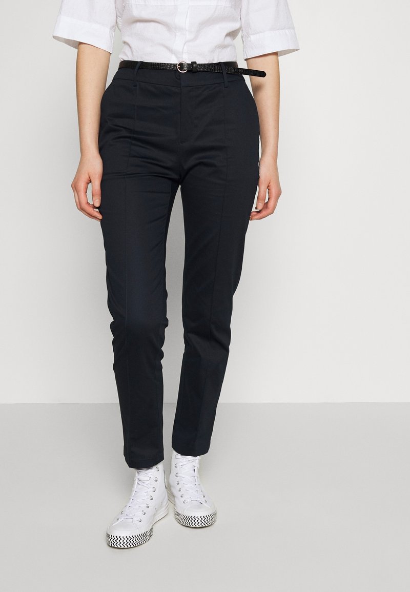 Personne portant un pantalon noir ajusté avec une ceinture noire fine, une chemise blanche à manches retroussées et des baskets montantes blanches.