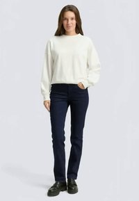 Naine seisab kotiga, seljas valge sviiter, tumesinised teksad ja mustad kingad, lihtsa tausta ees.