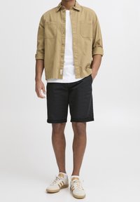 Camicia button-up beige con due tasche sul petto, T-shirt bianca sotto, pantaloni corti neri e sneakers crema con accenti marroni.