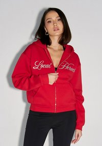 Roter Reißverschluss-Hoodie mit silberner, gestickter "Local Heroes"-Schrift auf der Brust, ausgestattet mit einer Fronttasche und einer lockeren Passform. Schwarze Hosen werden getragen.