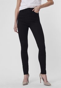 Black skinny jeans with a high waist, featuring a smooth denim texture, front pockets, and a fitted design, paired with beige high heels.