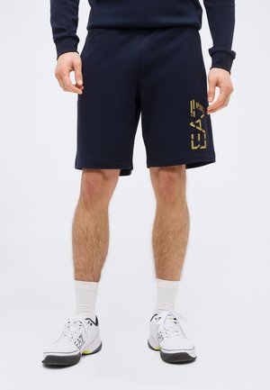 Férfi tengerészkék rövidnadrágban, bal lábán sárga "EA7" logóval, fehér zokniban és fehér sportcipőben, egyszínű háttér előtt állva.