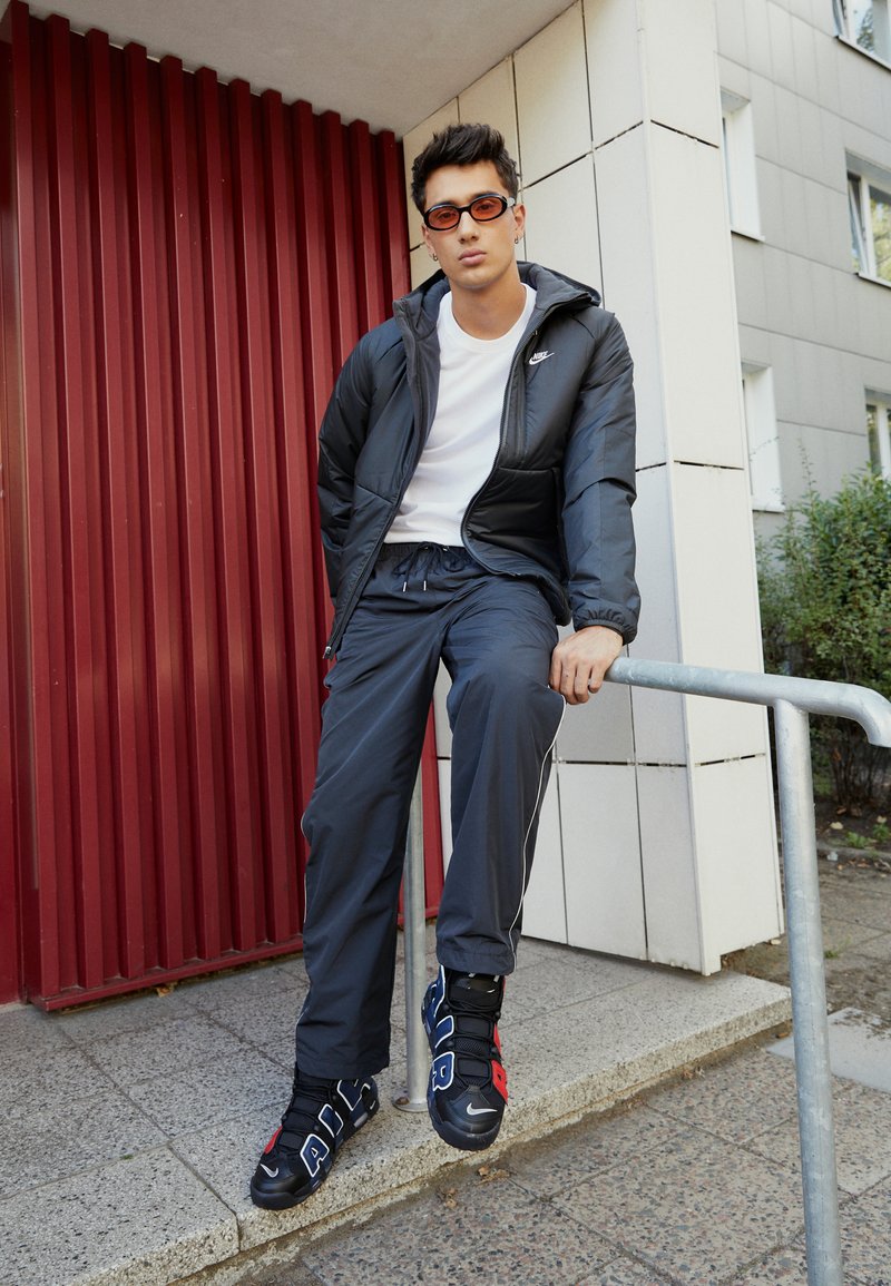 Black padded jacket with gray pants, a white t-shirt, and black shoes featuring red and blue accents, sitting on a metal railing.