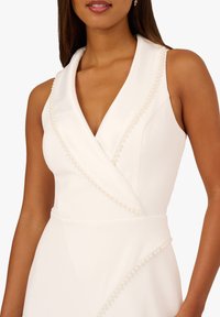 Bílé šaty bez rukávů s hlubokým výstřihem do V, zdobené perličkami podél výstřihu a diagonálního švu, vyrobené z hladké, strukturované látky.