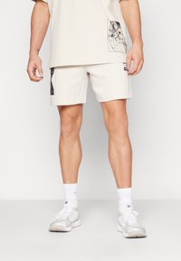 Calções de algodão bege claro com estampas gráficas. Textura suave, cintura elástica, corte reto. Combinados com meias atléticas brancas e sapatos.