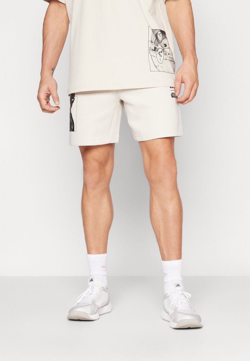 Beige bomullsshorts med sidografiktryck, med elastisk midja. I kombination med en matchande oversized skjorta och vita sneakers.