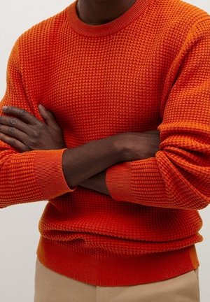 Mann mit dunkler Haut trägt einen strukturierten, leuchtend orangefarbenen Pullover, die Arme vor der Brust verschränkt, darunter sind beige Hosen sichtbar.