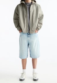 Veste à capuche gris clair avec des poignets élastiques, portée sur une chemise grise. Shorts en denim bleu à la coupe décontractée. Chaussures blanches avec des accents gris.