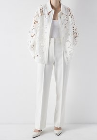 White open-front blouse with floral lace overlay, long sleeves, paired with high-waisted white trousers, and pointed-toe white heels.