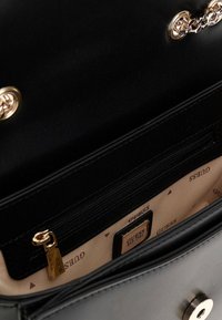 Bolso de cuero negro con acentos de cadena dorada, compartimento con cremallera y un interior estampado con la marca "Guess".